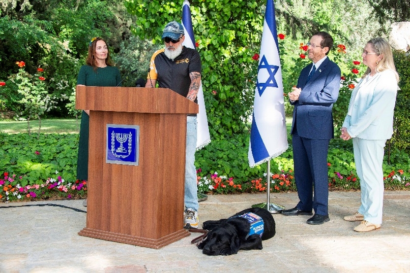 Benzi is pictured speaking at a podium with his wife Irit, his service dog Lugo, and Michal and President Yitzhak Herzog.